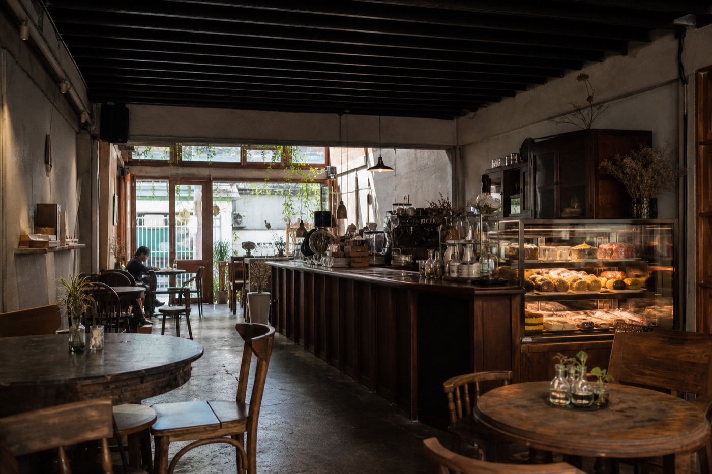 Illustrative dark-timber Bangkok cafe interior with a pastry display and tables near a leafy entrance